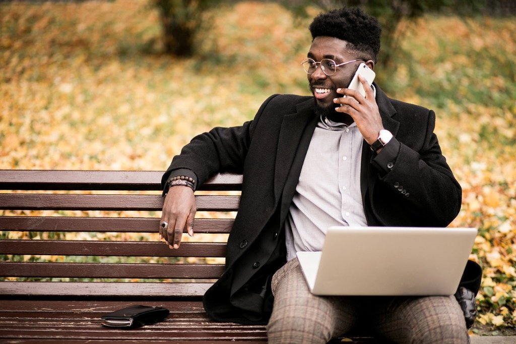 A phot of a user researcher sitting in the park whilst on the phone with his laptop on his knee.